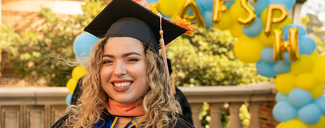 woman smiling in graduation cap and gown with balloons that say FSPH in the background