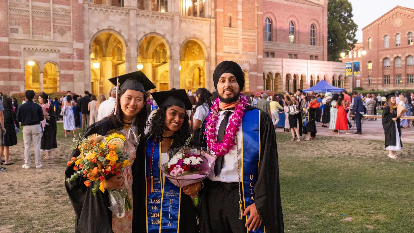 Three UCLA graduates in caps, gowns, and blue-and-gold stoles pose with bouquets and a flower lei outside of Royce Hall after commencement.